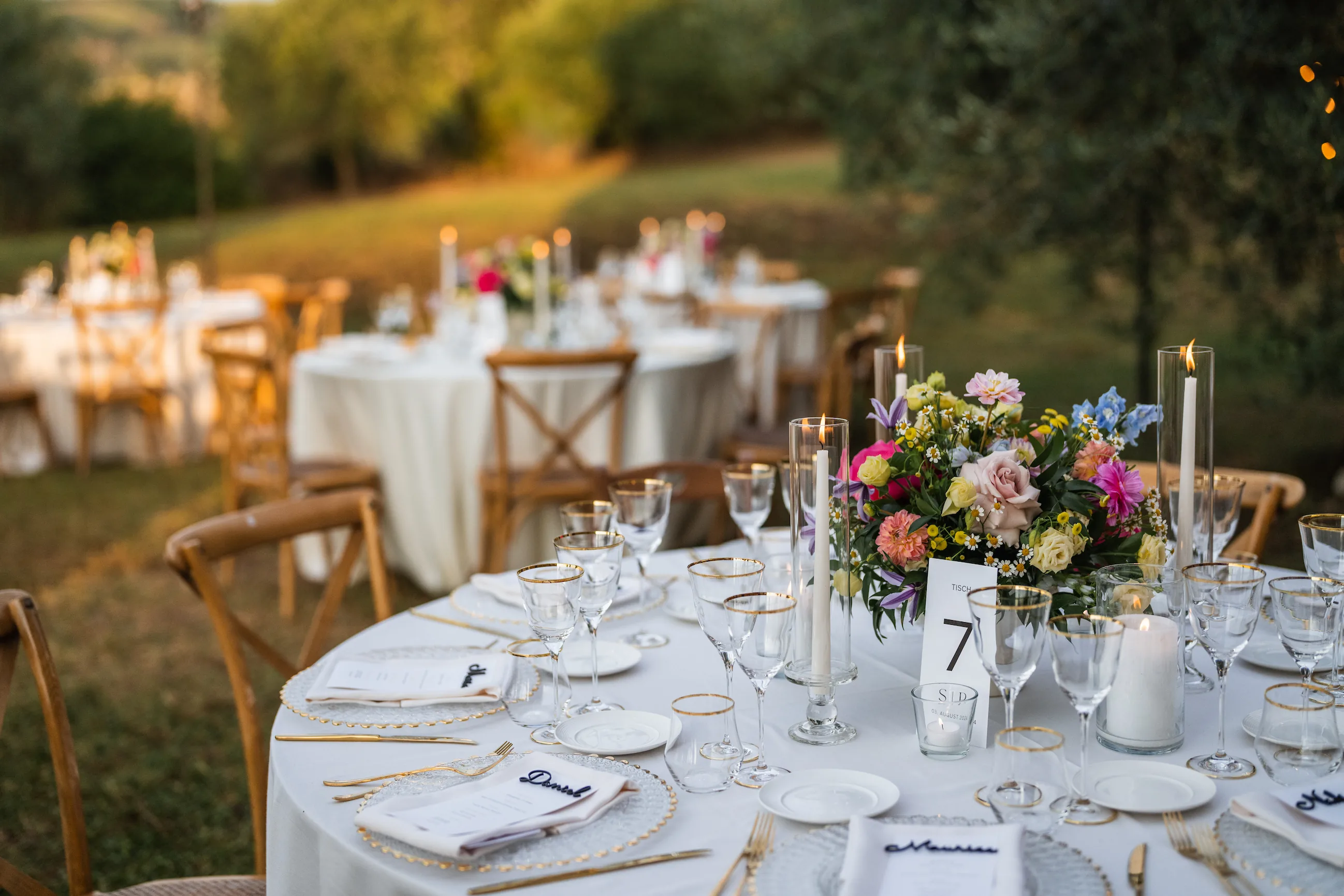 Runder Hochzeitstisch im Olivenhain bei Abendsonne mit bunten Blumen, Kerzen, Gläsern mit Goldrand und Namenskarten